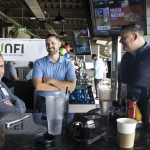 Three men stand and sit around a table at an indoor sports venue, smiling and talking. Drinks, condiments, and phones are on the table. Multiple screens and signs, including Fiesta Nachos, are visible in the background.
