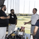 Four people stand and talk near a table with snacks and drinks at a golf driving range. Golf clubs are visible, and the range is seen through the netted windows in the background on a sunny day.