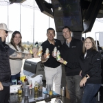 Five people stand and smile while raising bottled drinks together at an outdoor venue with golf bays in the background. A table with assorted drinks and glasses is in front of them.