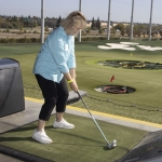 A woman wearing a blue top and black pants prepares to hit a golf ball at a driving range, aiming toward colorful target areas on the field. The Topgolf logo is visible on a nearby barrier.