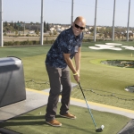 A man wearing sunglasses and a patterned shirt smiles while holding a golf club, standing on a driving range mat ready to hit a golf ball. The outdoor driving range is visible in the background.