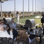 A group of people relax on couches at a Topgolf venue while one person stands swinging a golf club at the driving range. Drinks and snacks are on the table, with nets and targets visible outside.
