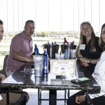 Four smiling adults sit around a high-top table at a golf driving range, with drinks and buckets of beer on the table. The outdoor golf bays are visible in the background through large windows.