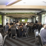 People sit at round tables in a modern, well-lit conference room, engaged in conversation. Large windows, screens on the walls, and event materials are visible. Some attendees are eating and chatting in a casual atmosphere.