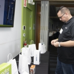 A man in a black shirt stands at a table with several white gift bags and small prizes. He is looking down at something in his hands. A partial presentation slide is visible on the wall behind him.