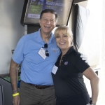 Two smiling adults, a man in a light blue polo and a woman in a black polo, stand close together indoors with name badges, posing for a photo near a golf simulator screen.