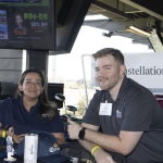 Two people sit at a table with Corona-branded items, smiling at the camera. Behind them are TV screens, a sign that reads Constellation Brands, and golf bays at a Topgolf venue.