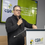 A man wearing glasses and a black pullover speaks into a microphone at a podium with the CGAEF logo. A screen behind him displays Welcome to the 2023 Top Shot Topgolf Tournament.