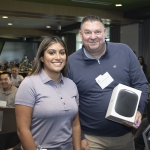 Two people smiling at an indoor event; one holds a boxed electronic device, possibly a prize. Several people sit at round tables in the background. The atmosphere is casual and friendly.
