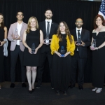 Seven people in formal attire holding awards, standing on a stage with a US flag behind them.