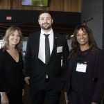 Three professionally dressed people smiling at an indoor event with tables in the background.