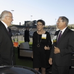 Three people in formal attire talk and smile at an outdoor event near a racetrack.