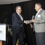 Two men in suits shake hands on stage as one receives an award at a formal event.