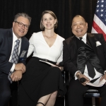 Three people in formal attire pose together, with an American flag in the background.