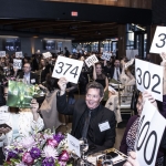 People at an auction event holding up numbered bidding cards in a crowded room.