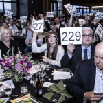 People at an auction raise numbered paddles while seated at decorated tables.