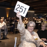 Woman at an auction holds up a paddle with the number 251 in a crowded room.