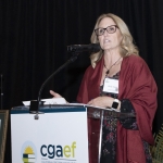 Woman speaking at a podium with CGAEF logo, wearing glasses and a red shawl.