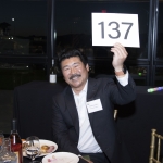 Smiling man in a suit holds up a sign with the number 137 at a formal event.