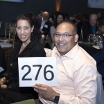 Smiling man holds a sign with 276 at a formal event, seated next to a woman.