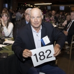 Man in a suit holding a sign with 182 at a formal event with a seated audience.