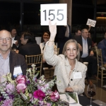 A woman holds up a card with 155 at a formal event, surrounded by people and flowers.