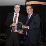 Two men in suits smiling and holding an award together at an indoor event.