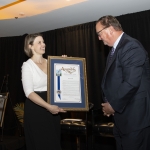 A woman presents a framed certificate to a man on a stage with a black curtain backdrop.
