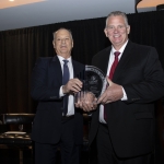 Two men in suits smiling and holding a glass award together on a stage with a black curtain.