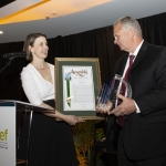 Two people exchange an award and framed certificate at a formal event on a stage.