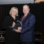 Two people in formal attire smiling and holding a glass award together on stage.