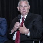 Man in suit and red tie speaking into a microphone against a dark background.