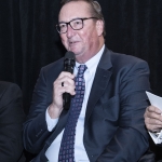 Man in a suit and glasses speaking into a microphone against a dark background.