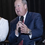 Man in a blue suit speaking into a microphone, seated against a dark curtain background.