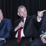 Three men in suits sit; the man in the middle speaks into a microphone and points to the right.