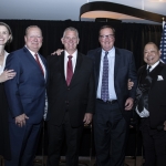 Five people in formal attire posing together indoors in front of a black curtain and a U.S. flag.